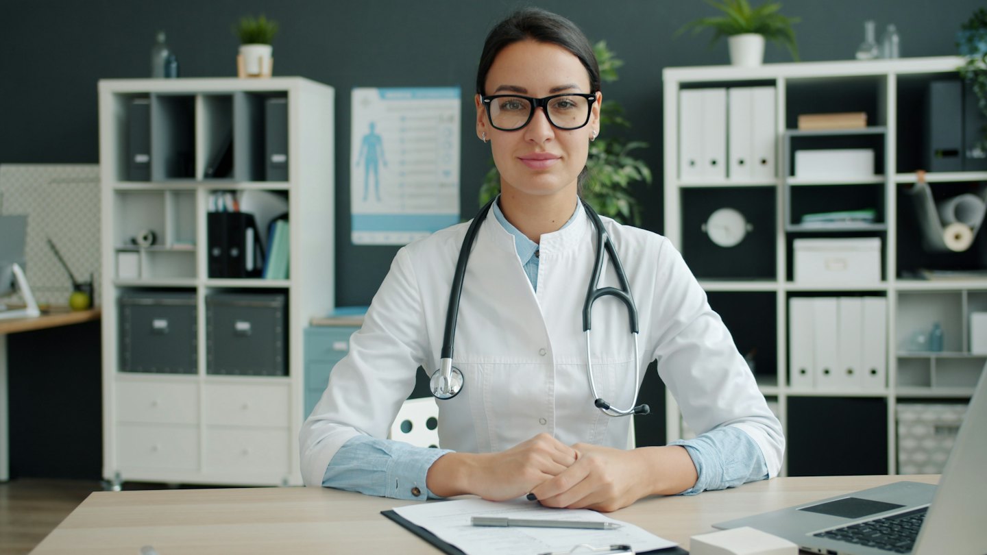 Doctor sits at a wooden desk with a laptop and clipboard.